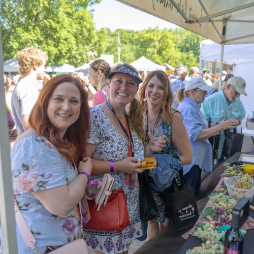 Wine festival attendees smiling near a vendor table with grapes and wine bottles in Downtown Excelsior Springs