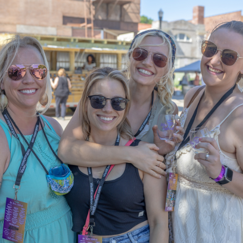 Friends wearing sunglasses holding wine glasses and smiling at an outdoor wine festival event