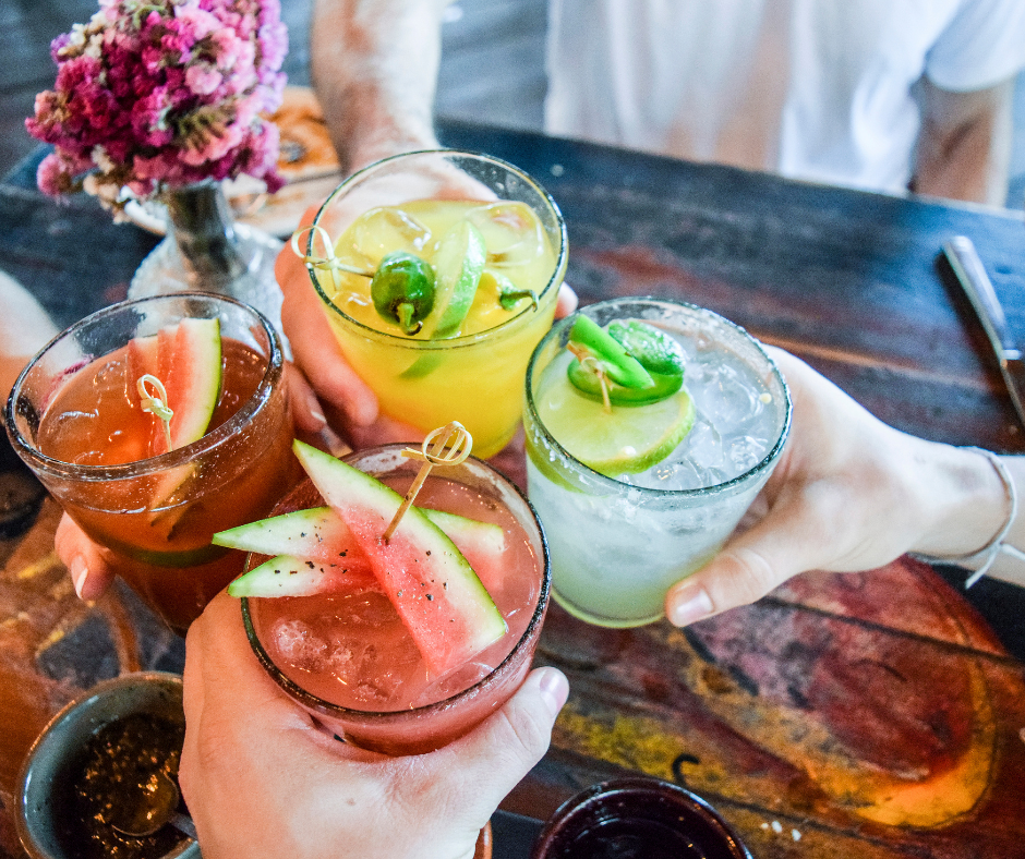 Four colorful cocktails raised in a toast over a rustic wooden table, garnished with watermelon slices, lime wedges, and jalapeño peppers.