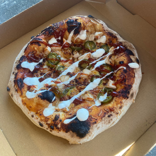 Wood-fired pizza with jalapeños, chicken, barbecue sauce, and white drizzle in a takeout box