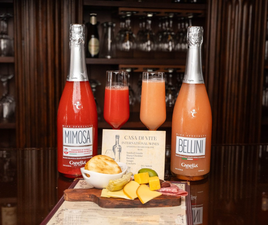 Charcuterie board with cheeses, crackers, and cured meats displayed alongside bottles of Mimosa and Bellini sparkling wine cocktails and filled flutes.