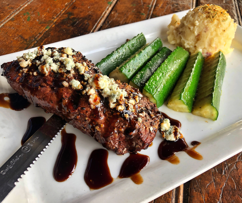 A plated steak topped with crumbled blue cheese and balsamic glaze is served alongside roasted zucchini and creamy mashed potatoes on a rustic wooden table at a downtown restaurant in Excelsior Springs. The image highlights the elevated dining experiences available during Second Weekends.