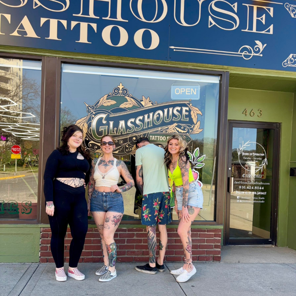 Group of tattoo artists posing outside Glasshouse Tattoo Studio storefront with large window signage in downtown Excelsior Springs.