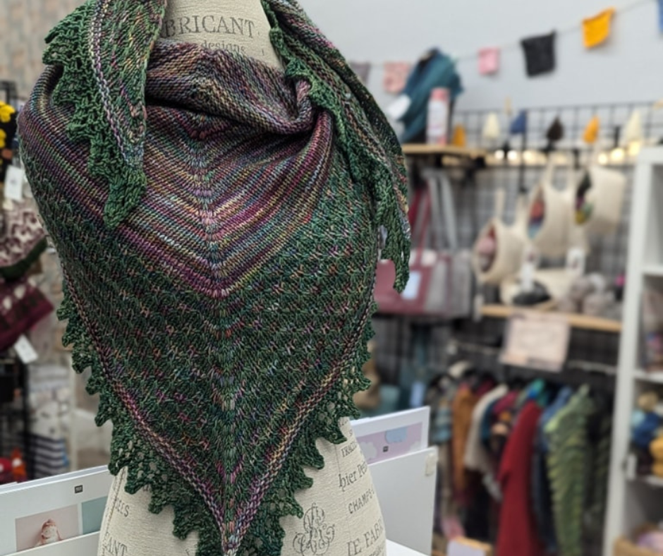 Hand-knit triangular shawl in shades of green, purple, and pink displayed on a dress form inside a fiber arts and yarn shop.