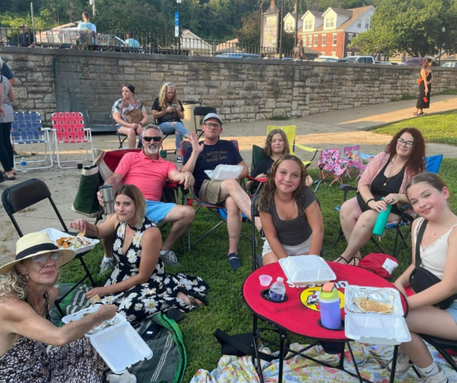 Families and friends gather on the lawn in Downtown Excelsior Springs with lawn chairs, picnic blankets, and takeout food during an outdoor community event. Attendees relax and enjoy the summer evening atmosphere at Lyrics on the Lawn during Second Weekends.