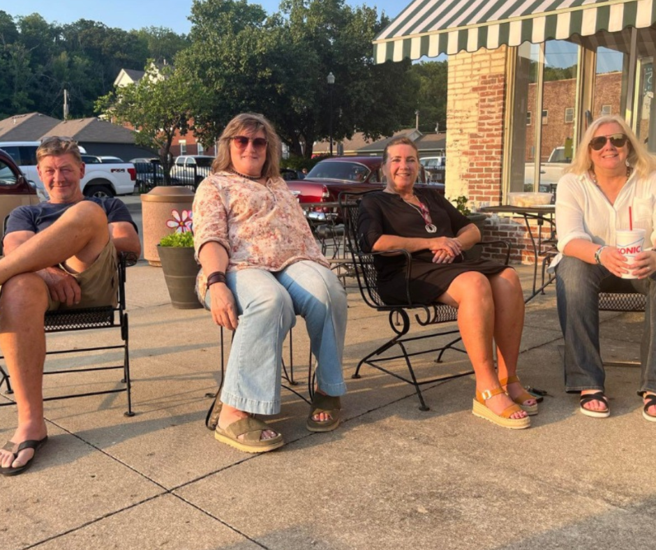 Four adults relax in lawn chairs outside a downtown storefront during an evening community event in Downtown Excelsior Springs. The group enjoys warm weather, conversation, and the laid-back atmosphere of Lyrics on the Lawn during Second Weekends.
