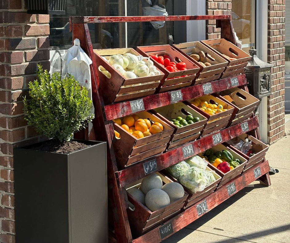 Outdoor produce stand featuring wooden crates filled with fresh fruits and vegetables including tomatoes, peppers, melons, citrus, and onions.