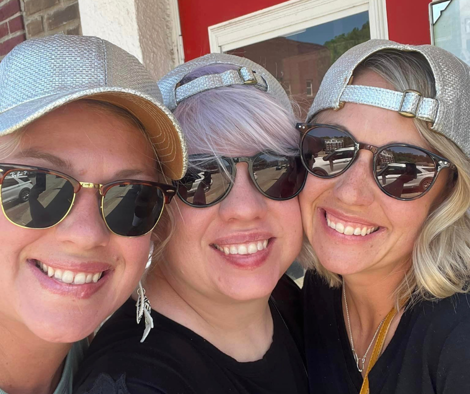 Three women wearing silver hats and sunglasses smile together for a selfie in Downtown Excelsior Springs during a sunny Second Weekends outing. The group appears to be enjoying a fun day of shopping, dining, and exploring downtown.
