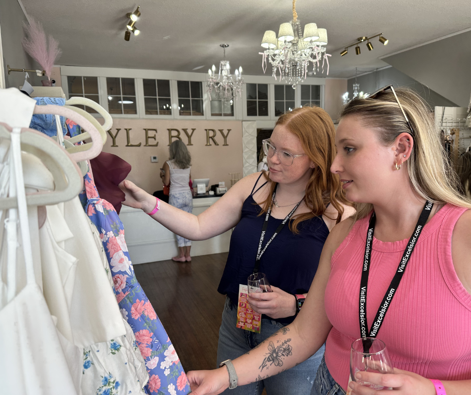Two women browse dresses inside Style By Ry boutique in Downtown Excelsior Springs while holding wine glasses during a Shop Local Late event. The image captures the shopping, social atmosphere, and local experiences featured during Second Weekends.