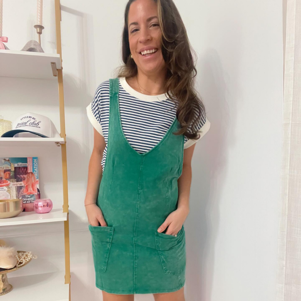 Woman modeling a green corduroy jumper dress over a striped shirt inside Style By Ry boutique in downtown Excelsior Springs.