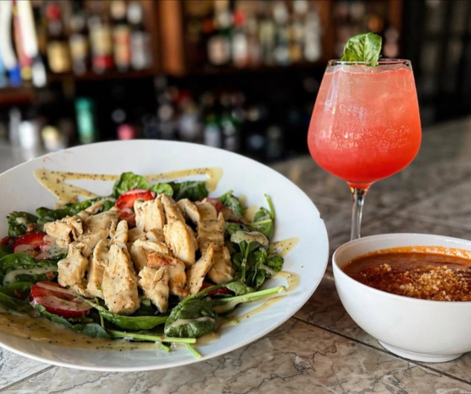 Fresh salad topped with grilled chicken served alongside a bowl of soup and a bright pink specialty cocktail at a restaurant table.