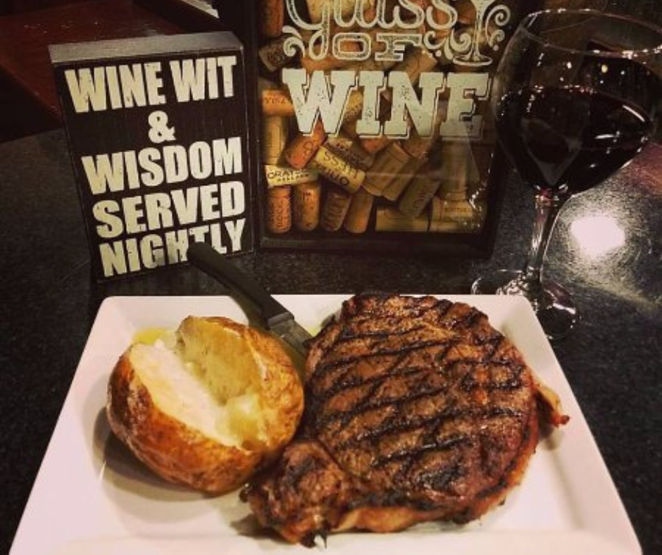 Grilled steak and baked potato plated beside a glass of red wine, with wine-themed décor displayed in the background.