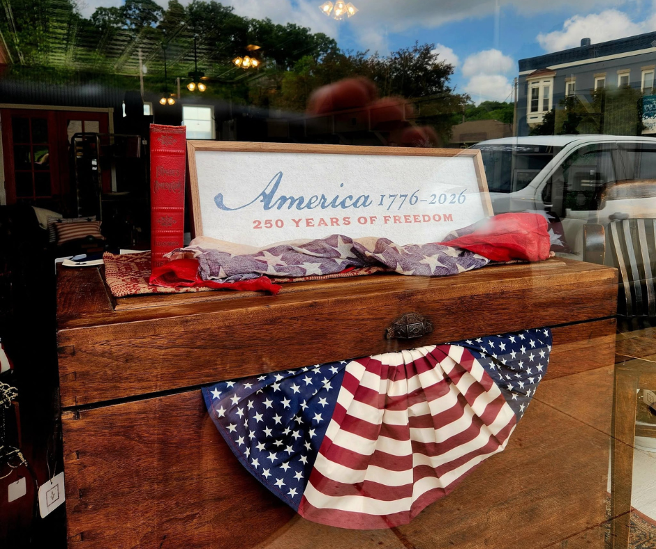 Patriotic storefront display featuring a vintage wooden chest decorated with red, white, and blue bunting. Resting on top is a sign reading “America 1776–2026: 250 Years of Freedom,” alongside a red antique-style book and a folded American flag-themed cloth. Reflections of downtown buildings, parked vehicles, and a blue sky with clouds are visible in the shop window.
