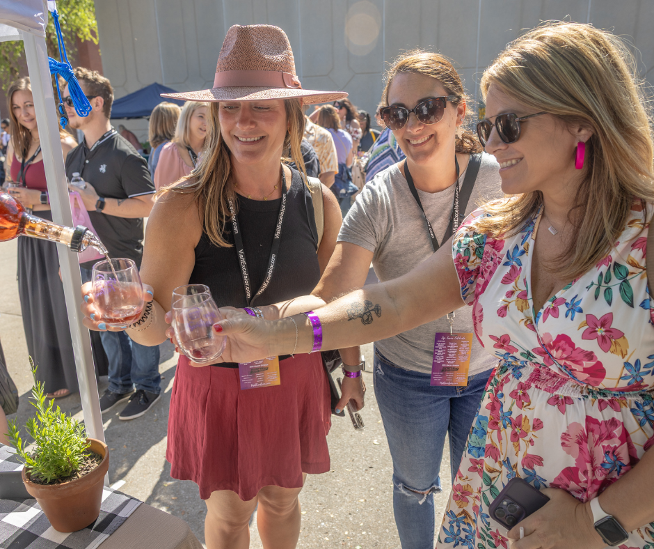 Three women smile and hold out wine glasses while sampling rosé wine during a lively outdoor festival in Downtown Excelsior Springs. Attendees wear tasting lanyards and gather among vendor booths and crowds in the sunny downtown street atmosphere.