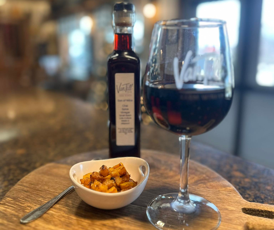 A glass of red wine and a bottle of balsamic vinegar sit on a wooden serving board beside a small bowl of roasted appetizer bites inside a winery tasting room in Downtown Excelsior Springs. The image highlights the wine, food pairings, and upscale tasting experiences available during Second Weekends.