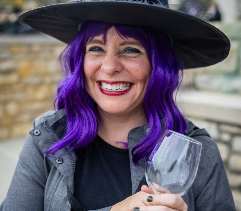 Smiling attendee wearing a witch hat and purple wig holds a wine glass.