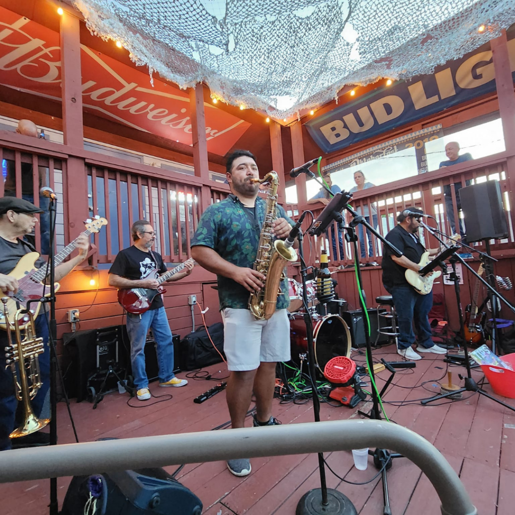 Members of All Star Band KC perform live on an outdoor stage during a summer concert, featuring saxophone, guitars, drums, and brass instruments under string lights at a local music venue.
