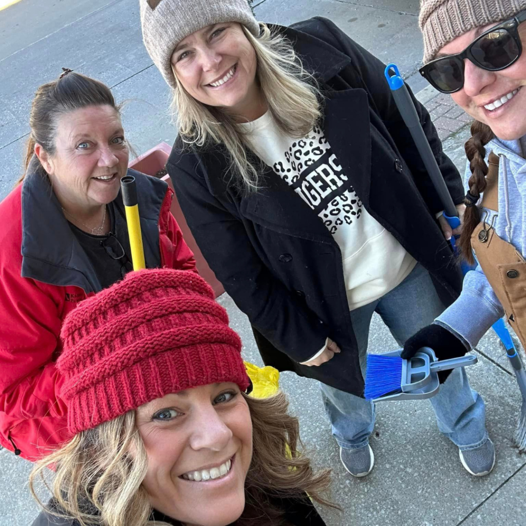 Carol Baxter and fellow Downtown Excelsior Partnership volunteers smile while participating in a downtown cleanup day, holding brooms and tools along a sidewalk in Downtown Excelsior Springs.