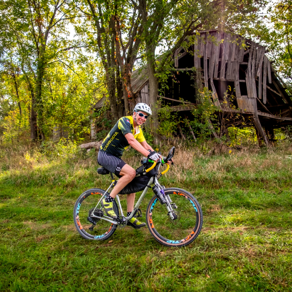A cyclist rides along a gravel route past a historic barn during the Opal Wapoo Gravel Experience, showcasing the scenic rural landscapes surrounding Excelsior Springs.
