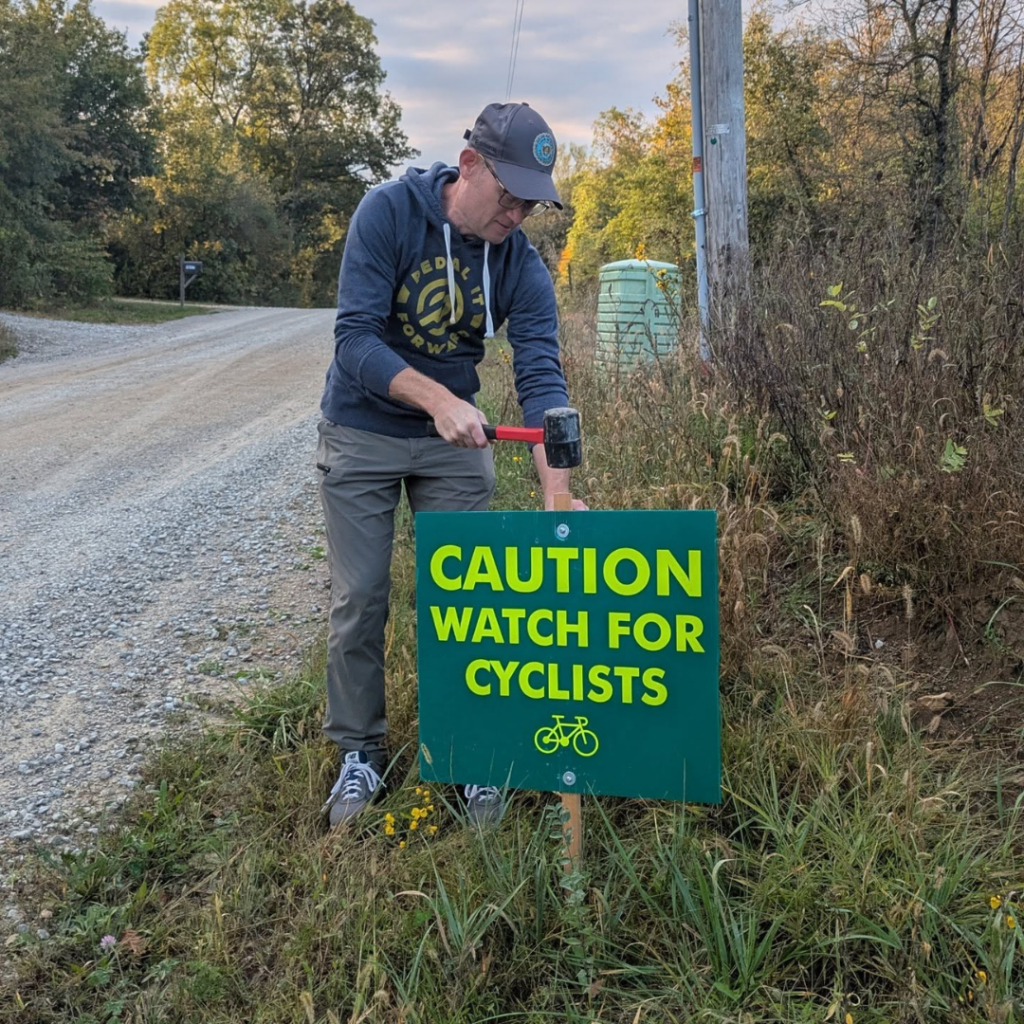 A volunteer installs a roadside caution sign before the Opal Wapoo Gravel Experience to help ensure cyclist safety along rural gravel roads.