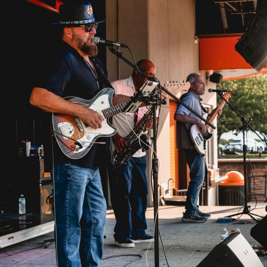 Members of KC Ruckus Band perform live on an outdoor stage, featuring guitar, saxophone, bass, and vocals during a summer concert.