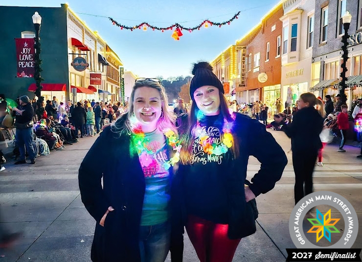 Two women smile in a downtown street filled with people and holiday lights during Christmas Kickoff.