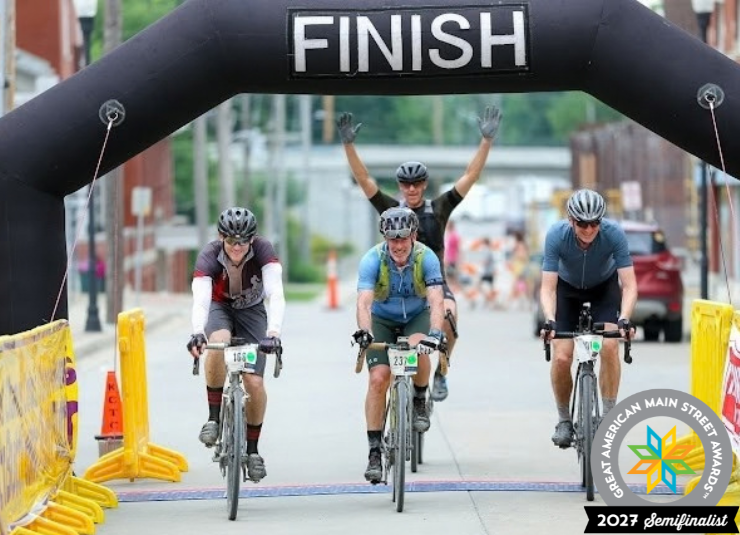 Four cyclists cross the finish line of a downtown bike ride, with one rider raising both arms.