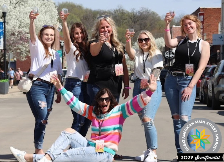 Five women raise tasting glasses on a downtown street while a sixth sits in front with her arms raised.